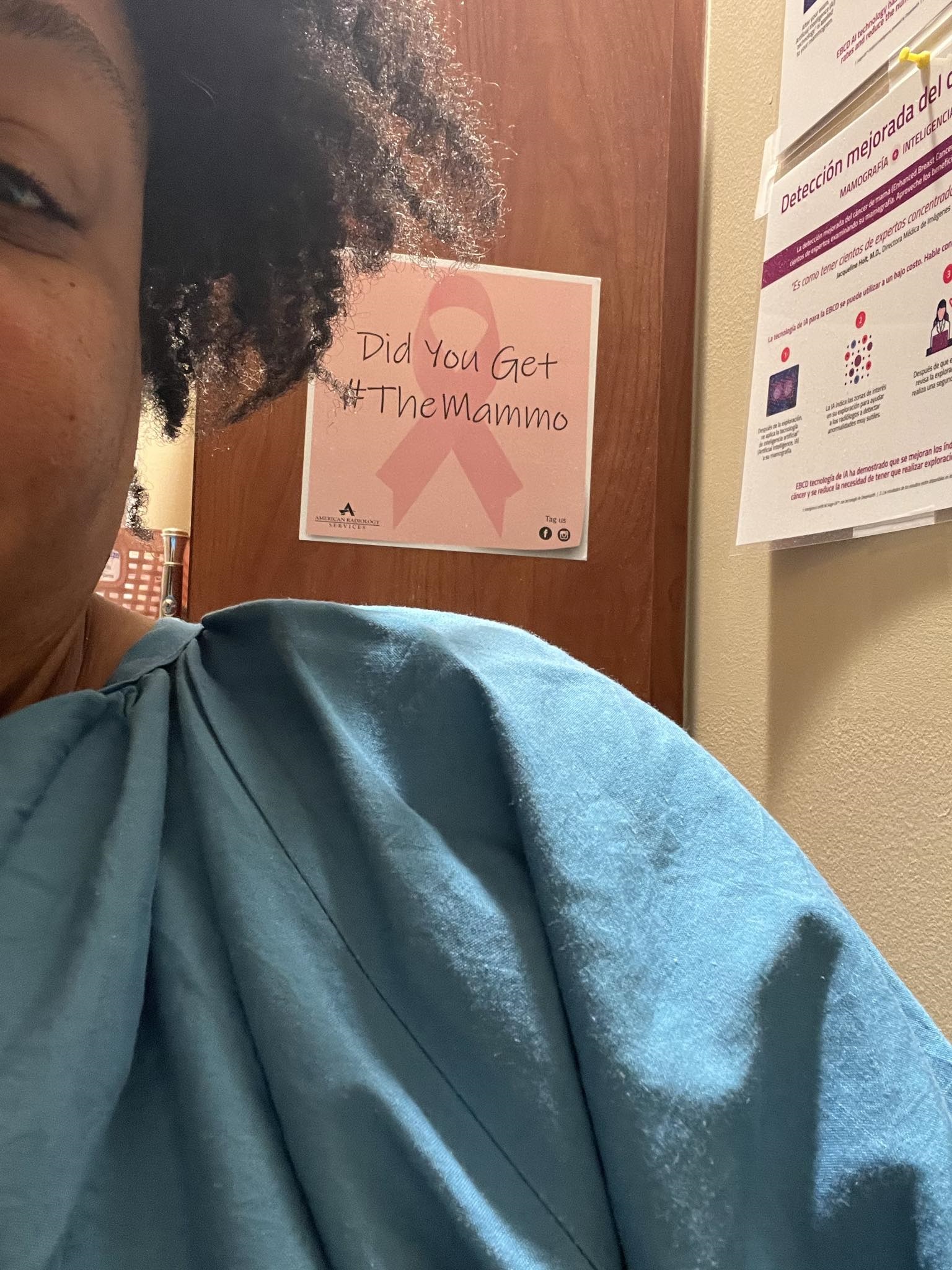 Tamika Felder in a hospital gown at her radiologist's office, ready to get her mammogram. She stands in front of a wall with a sign that says, "Did You Get #TheMammo"?