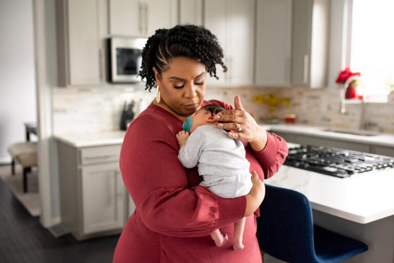 Tamika Felder, wearing a rose-colored top, holds her newborn son, Chayton, who has a pacifier in his mouth. They are in a light-filled kitchen. Tamika reflects on missing her preventative screenings last year due to the distractions of new motherhood.
