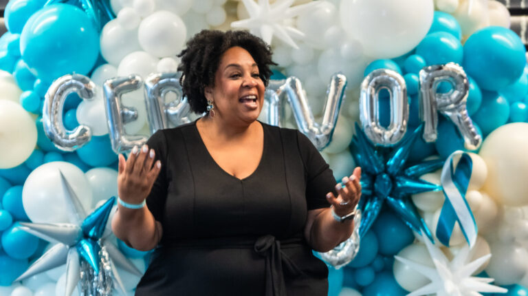 Tamika Felder, in a black jumpsuit, speaks to a group of Cervivors, cervical cancer patients, and survivors, with blue and teal balloons behind her. Cervivor is spelled out in silver mylar balloons.