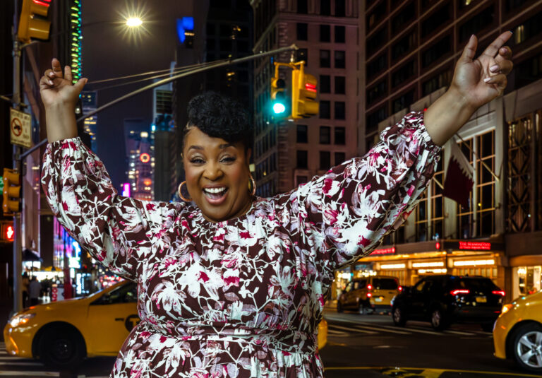 Tamika Felder in New York City, wearing a floral dress and smiling joyfully with arms raised.