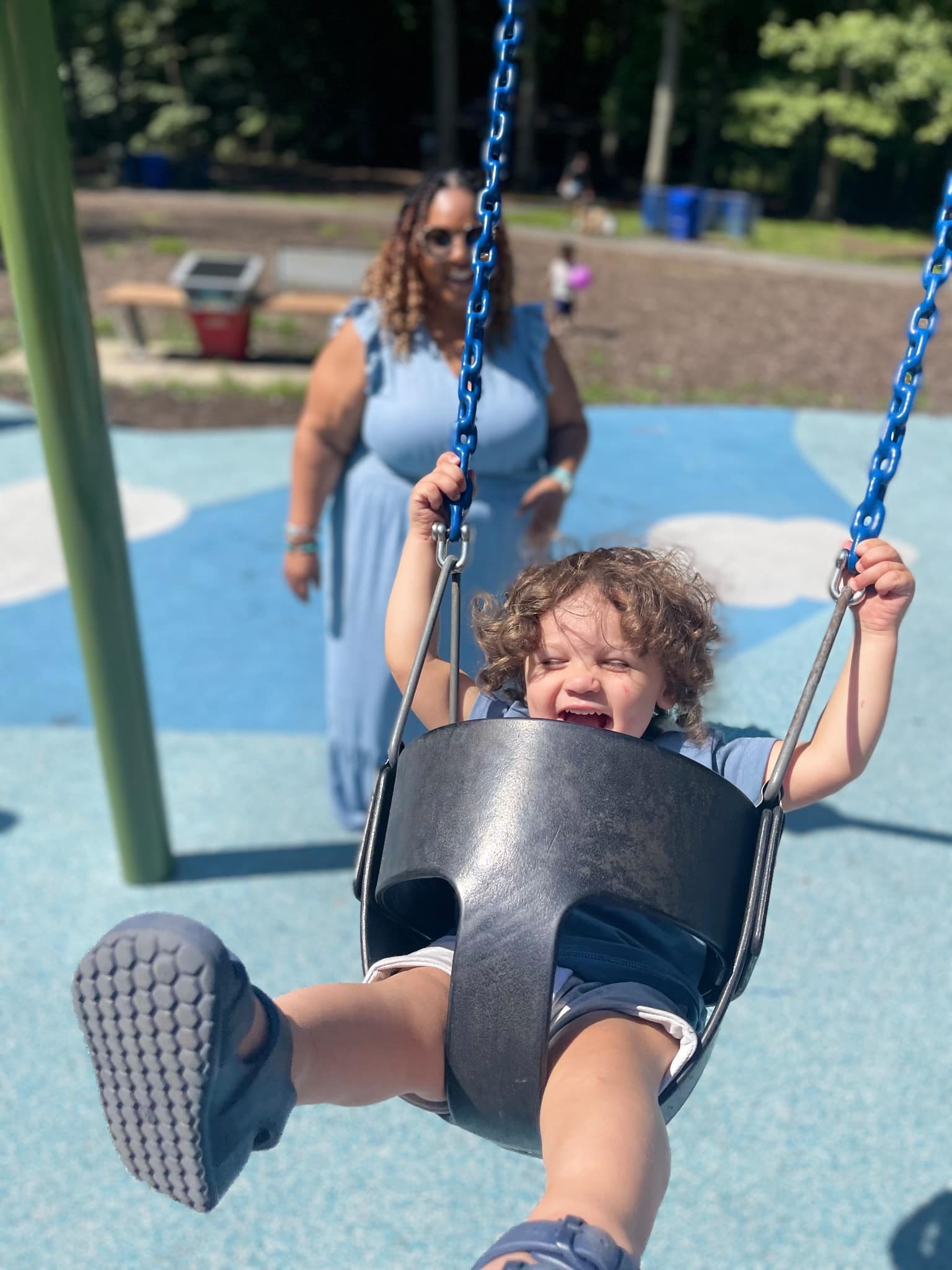 Tamika Felder pushes her toddler son, Chayton, on a swing at a playground on a sunny day. Chayton, who is under 2, is smiling with joy as he swings, while Tamika stands behind him in a blue dress, enjoying the moment.