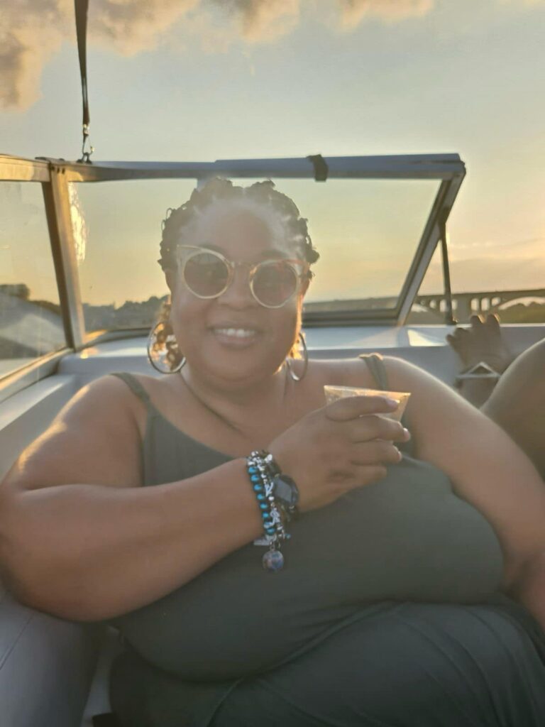 Tamika Felder, global cancer advocate and motivational speaker, enjoys a summer boat ride on the Potomac River. She is smiling, wearing sunglasses, and holding a drink, with the sun setting behind her, creating a warm, golden glow.