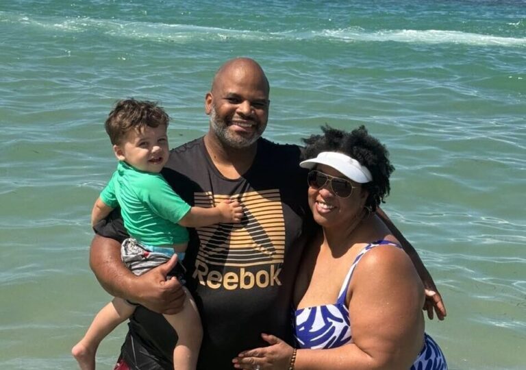 Tamika Felder and her husband, Rocky, enjoying a beach day in Puerto Rico with their two-year-old son, Chayton. Rocky is holding Chayton, and the family is dressed in beach attire, standing in front of a scenic backdrop with sand and ocean waves under a bright blue sky.