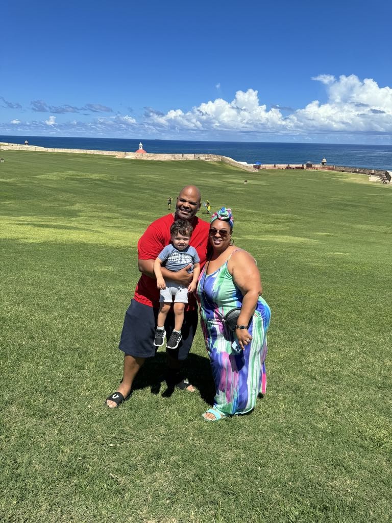 Tamika Felder, her husband Rocky, and their young son Chayton pose together on a lush green field with a stunning ocean view in the background during their beach vacation to Puerto Rico.