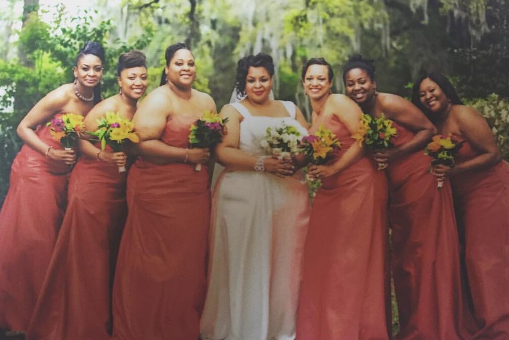 Tamika Felder on her wedding day in South Carolina, wearing a long white gown and holding flowers, surrounded by her bridesmaids, including best friend Quan Myers. The bridesmaids wear long burnt orange dresses, with a stunning backdrop of big trees draped in Spanish moss.