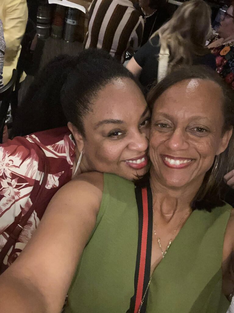 Best friends Tamika Felder and Quan Myers smile at the camera in a selfie. Quan holds the camera while wearing a green shirt, and Tamika stands behind her in a red and white shirt with a long ponytail.