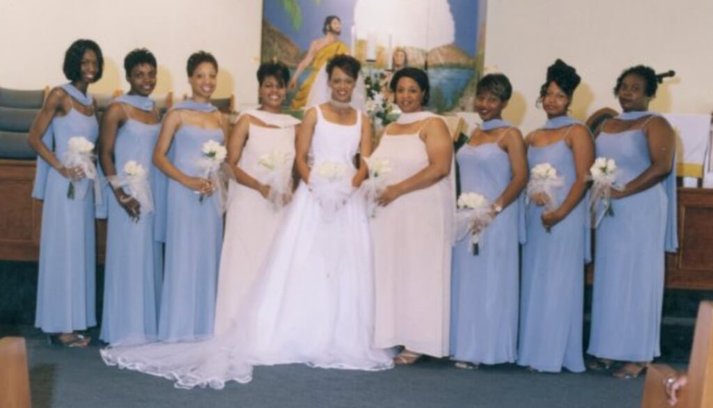 Quan Myers on her wedding day 25 years ago, surrounded by her bridesmaids, including Tamika Felder. Quan is wearing a long white wedding gown, and the bridesmaids are in long light blue dresses, all smiling for the camera.