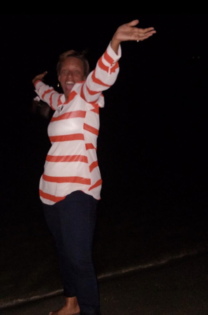 Quan Myers joyfully throws her arms out with a big smile on her face while outdoors at night, wearing an orange and white striped shirt and dark blue pants.