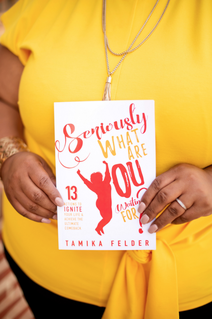 A person wearing a bright yellow top holds Seriously, What Are You Waiting For? 13 Actions to Ignite Your Life & Achieve the Ultimate Comeback by Tamika Felder, with a striking red and yellow cover featuring a leaping figure, set against a soft outdoor backdrop.
