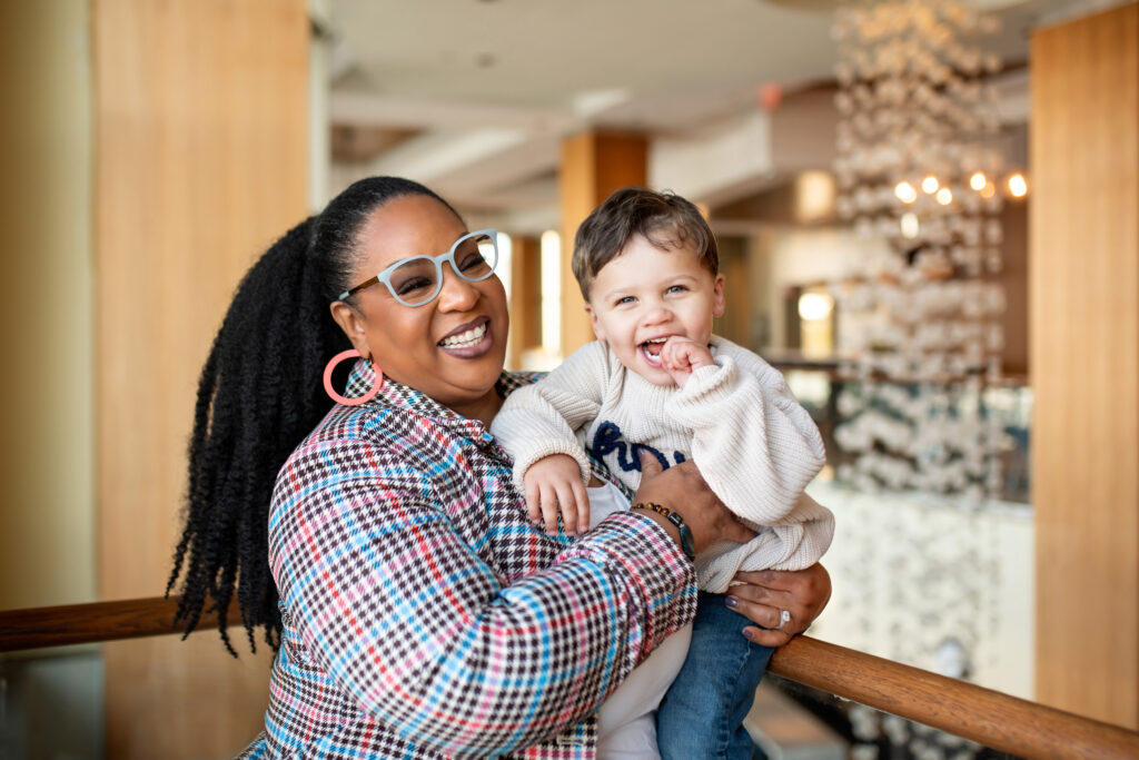 Tamika Felder, Cervivor founder, smiles warmly while holding her son Chayton, who grins happily. She is dressed in a vibrant plaid blazer and stands by a wooden railing in a cozy indoor space with wooden pillars and decorative elements in the background.