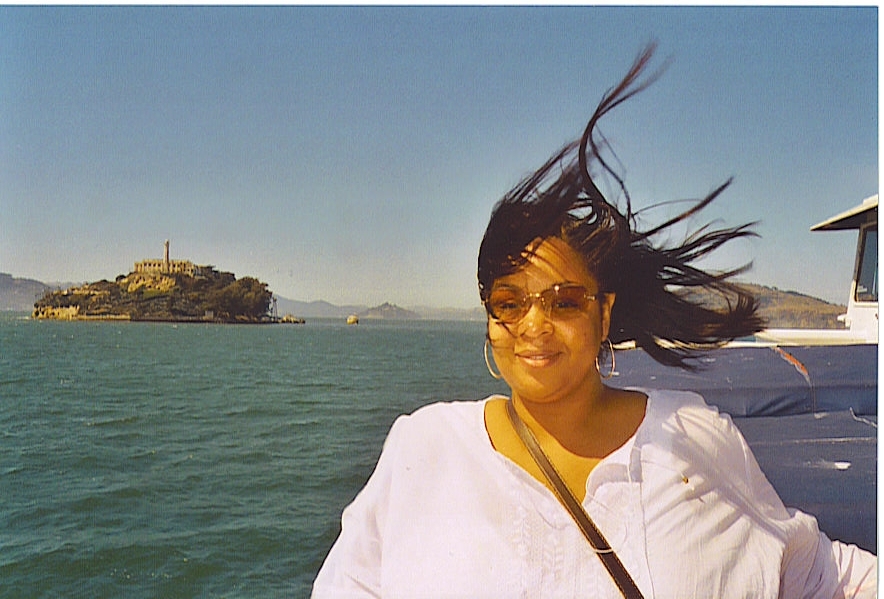 A young Tamika Felder smiles on a breezy boat ride with Alcatraz Island visible in the background. Her hair is windswept, and she’s wearing sunglasses and a white top, captured in a carefree moment before her cancer diagnosis.