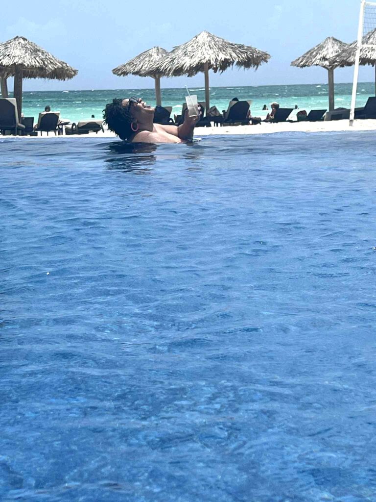 Tamika Felder floats happily in a pool holding a drink, wearing sunglasses, with thatched umbrellas, lounge chairs, and a turquoise ocean in the background under a clear sky.